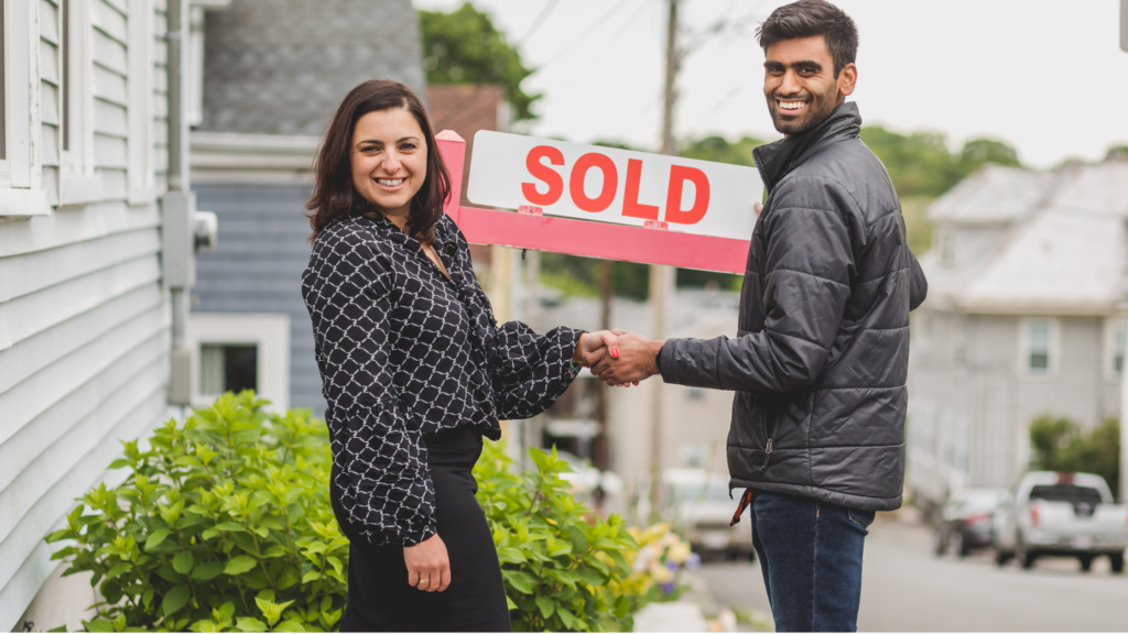 A man and woman shaking hands in front of a house with a sold sign, indicating a successful real estate transaction.