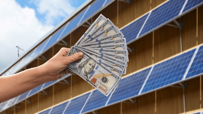 A hand holding a fan of hundred-dollar bills in front of a building covered in solar panels, debunking the myth that solar energy isn't profitable. forme solar