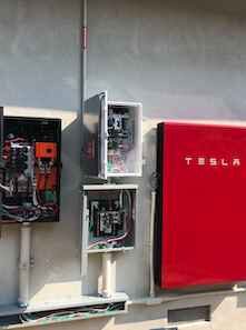 An electrical setup featuring an open circuit breaker box next to a closed red Tesla power unit mounted on a wall.