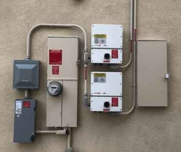 Two electrical boxes and utility meters mounted on an exterior wall with exposed pipes and conduits. Two electrical boxes and utility meters mounted on an exterior wall with exposed pipes and conduits.